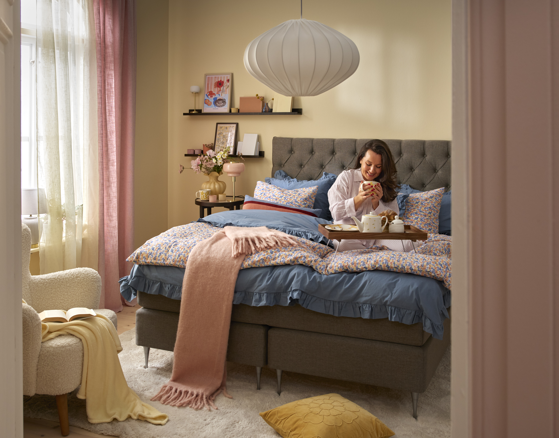 En kvinna äter frukost i Asperö kontinentalsäng i grått med blåa och blommiga sängkläder.