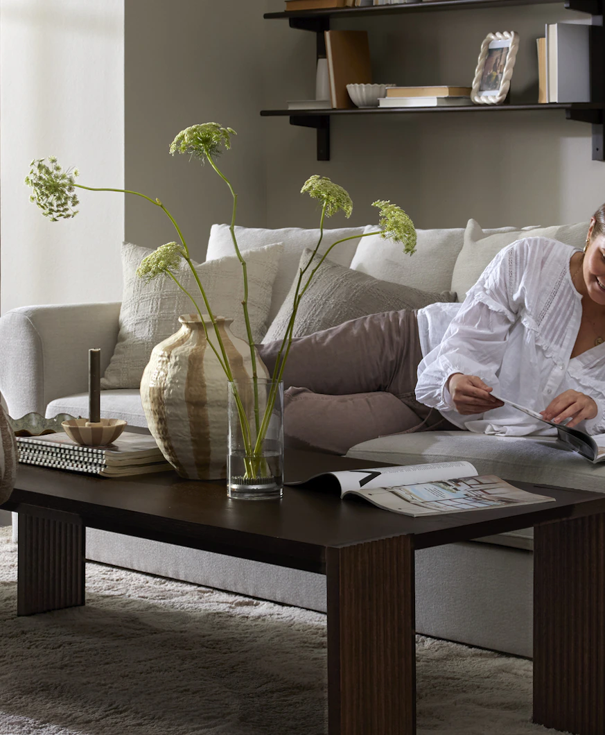 I ett grått vardagsrum ligger en kvinna i en ljusgrå soffa och bläddrar i ett magasin.