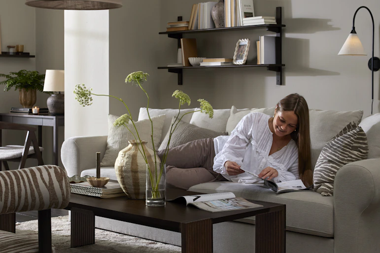 I ett grått vardagsrum ligger en kvinna i en ljusgrå soffa och bläddrar i ett magasin.