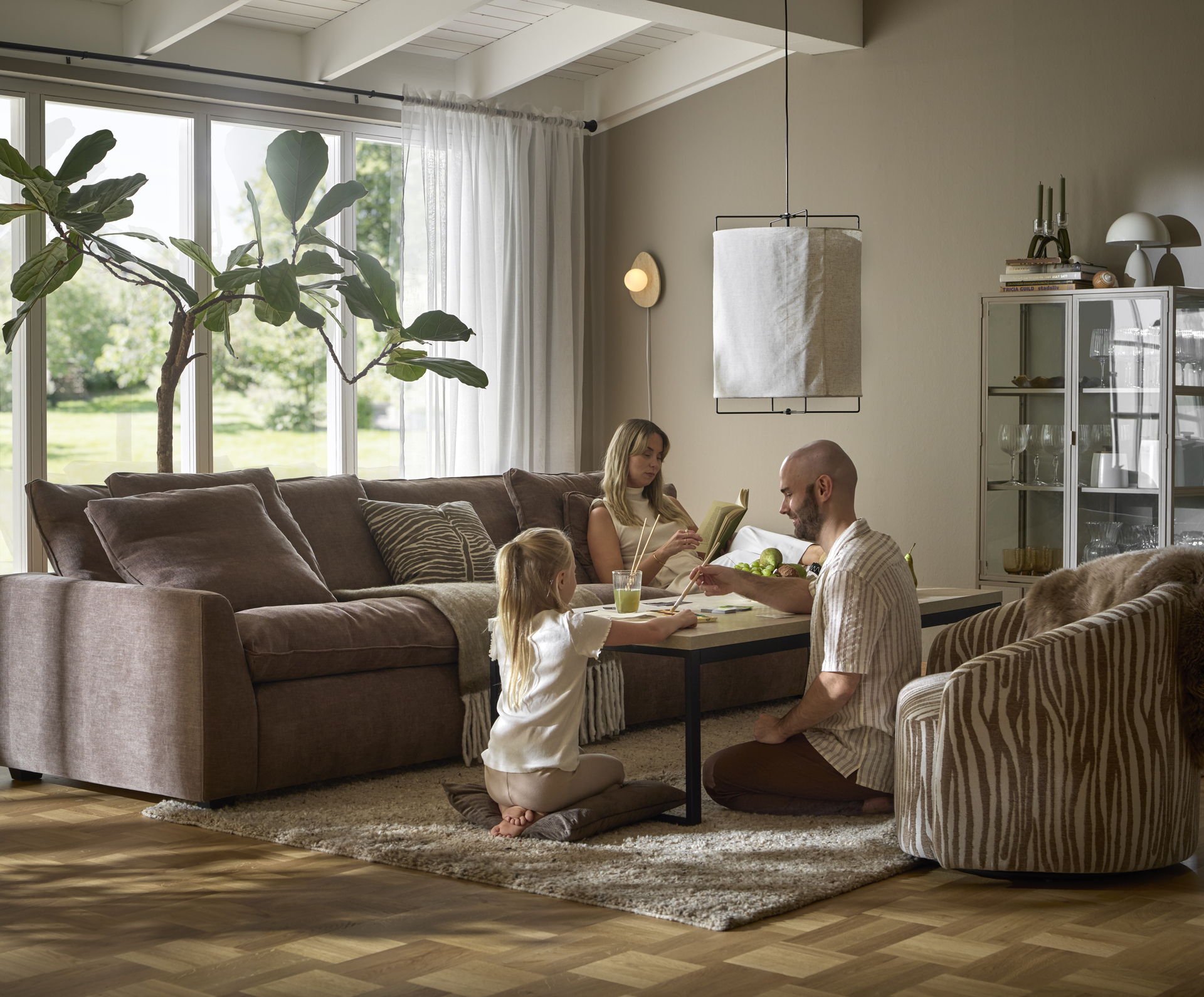 En kvinna sitter i en stor, brun soffa och läser, på andra sidan soffbordet sitter en man och ett barn och målar med vattenfärg