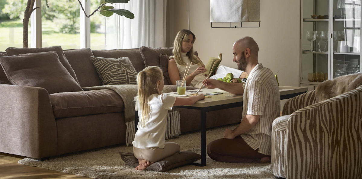 En kvinna sitter i en stor, brun soffa och läser, på andra sidan soffbordet sitter en man och ett barn och målar med vattenfärg