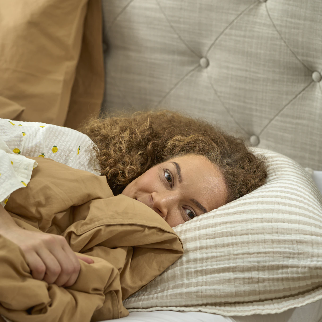 Närbild på ung kvinna i säng, ligger med huvudet på kudden och tittar in i kameran. 