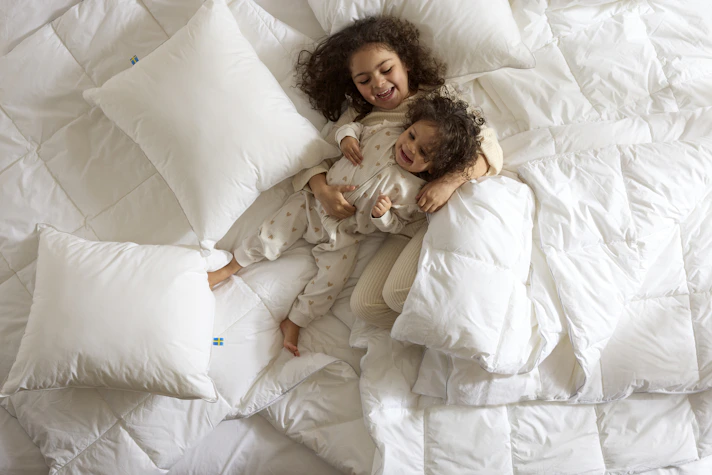 Två barn ligger och busar i en säng bland kuddar och täcken, foto taget direkt ovanifrån.