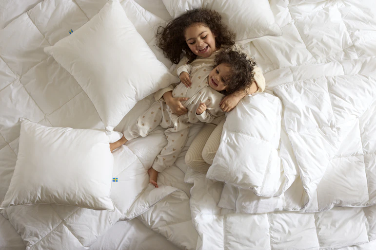 Två barn ligger och busar i en säng bland kuddar och täcken, foto taget direkt ovanifrån.