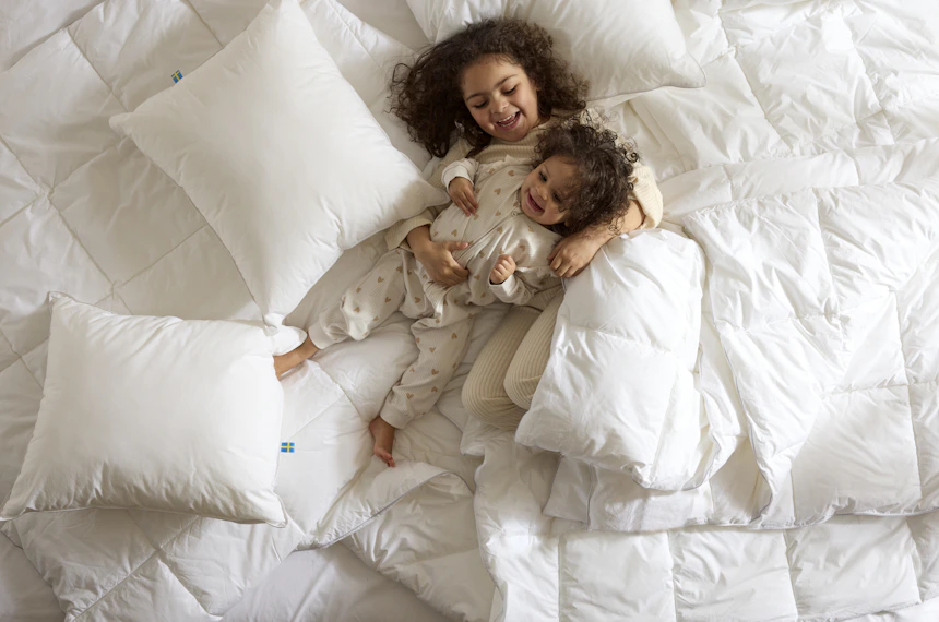 Två barn ligger och busar i en säng bland kuddar och täcken, foto taget direkt ovanifrån.