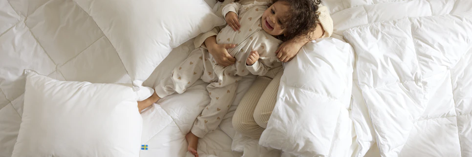 Två barn ligger och busar i en säng bland kuddar och täcken, foto taget direkt ovanifrån.