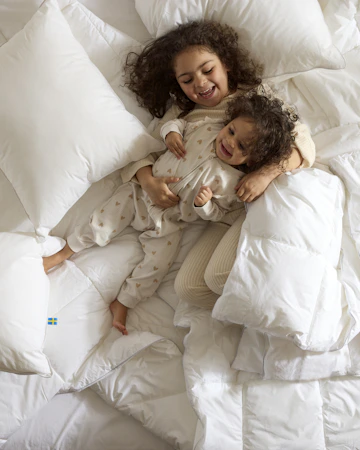 Två barn ligger och busar i en säng bland kuddar och täcken, foto taget direkt ovanifrån.