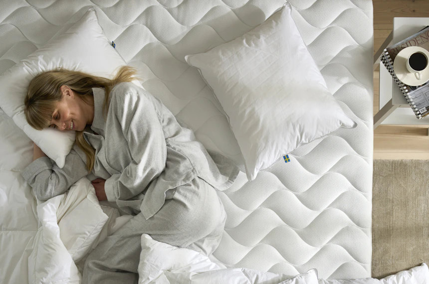 Kvinna ligger leendes på sidan i säng och gosar in sig i täcken och kuddar.