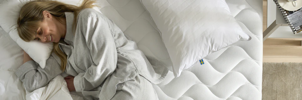 Kvinna ligger leendes på sidan i säng och gosar in sig i täcken och kuddar.