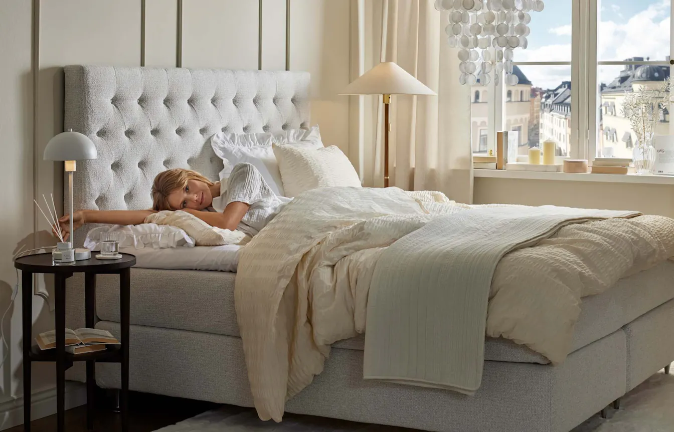 Kvinna ligger på sidan med utsträckt arm under huvudet i en säng i ett ljust sovrum, i ett fönster syns att det är fint väder ute.