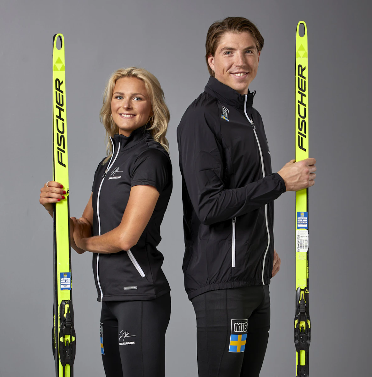 Längdskidåkarna Frida Karlsson och. William Poromaa rygg mot rygg med träningskläder och håller i skidor i en studiomiljö.