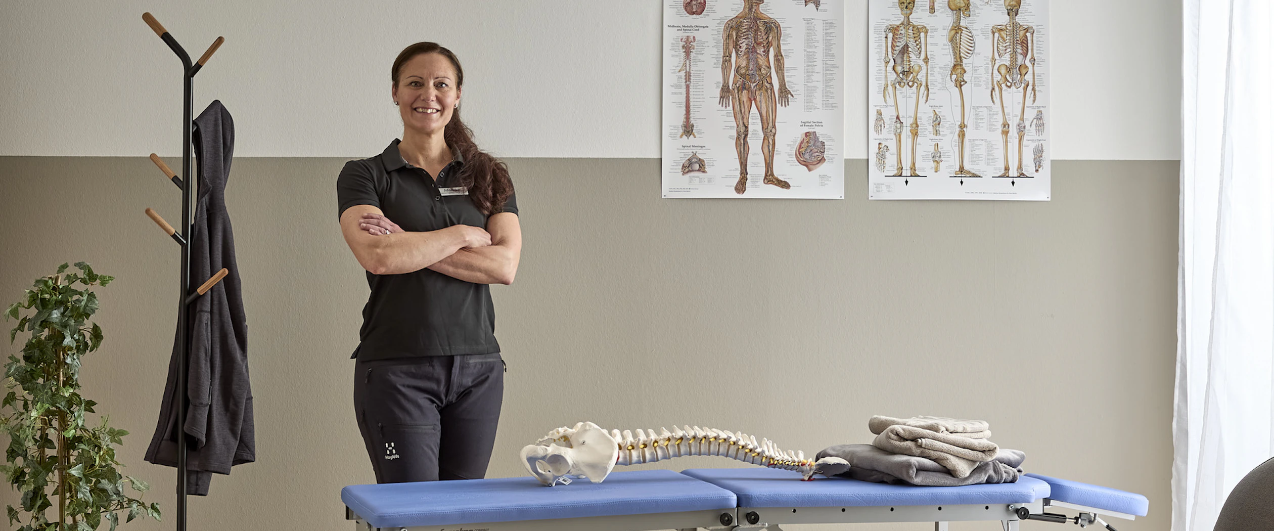 Monika Seth som är ergonom och fysioterapeut står i fysiomottagning bakom en brits med ergonomiska planscher på väggen bakom.