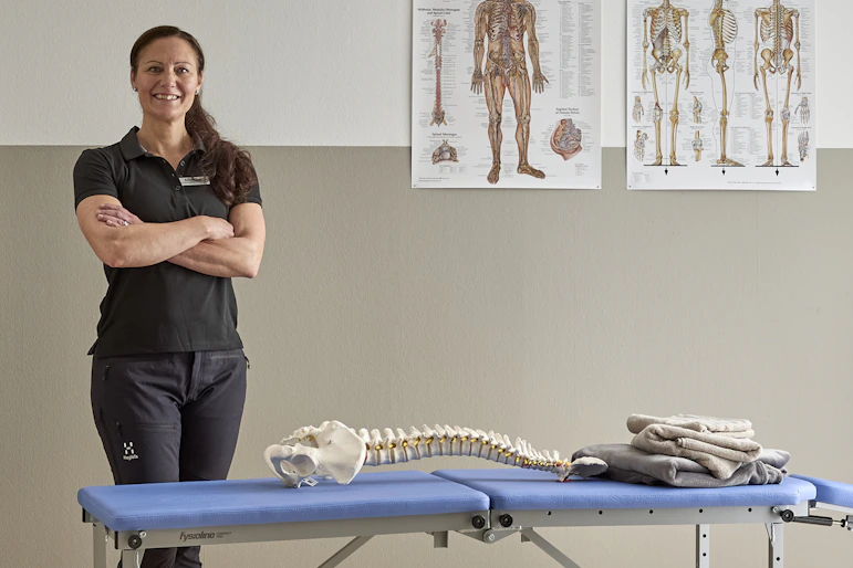 Monika Seth som är ergonom och fysioterapeut står i fysiomottagning bakom en brits med ergonomiska planscher på väggen bakom.