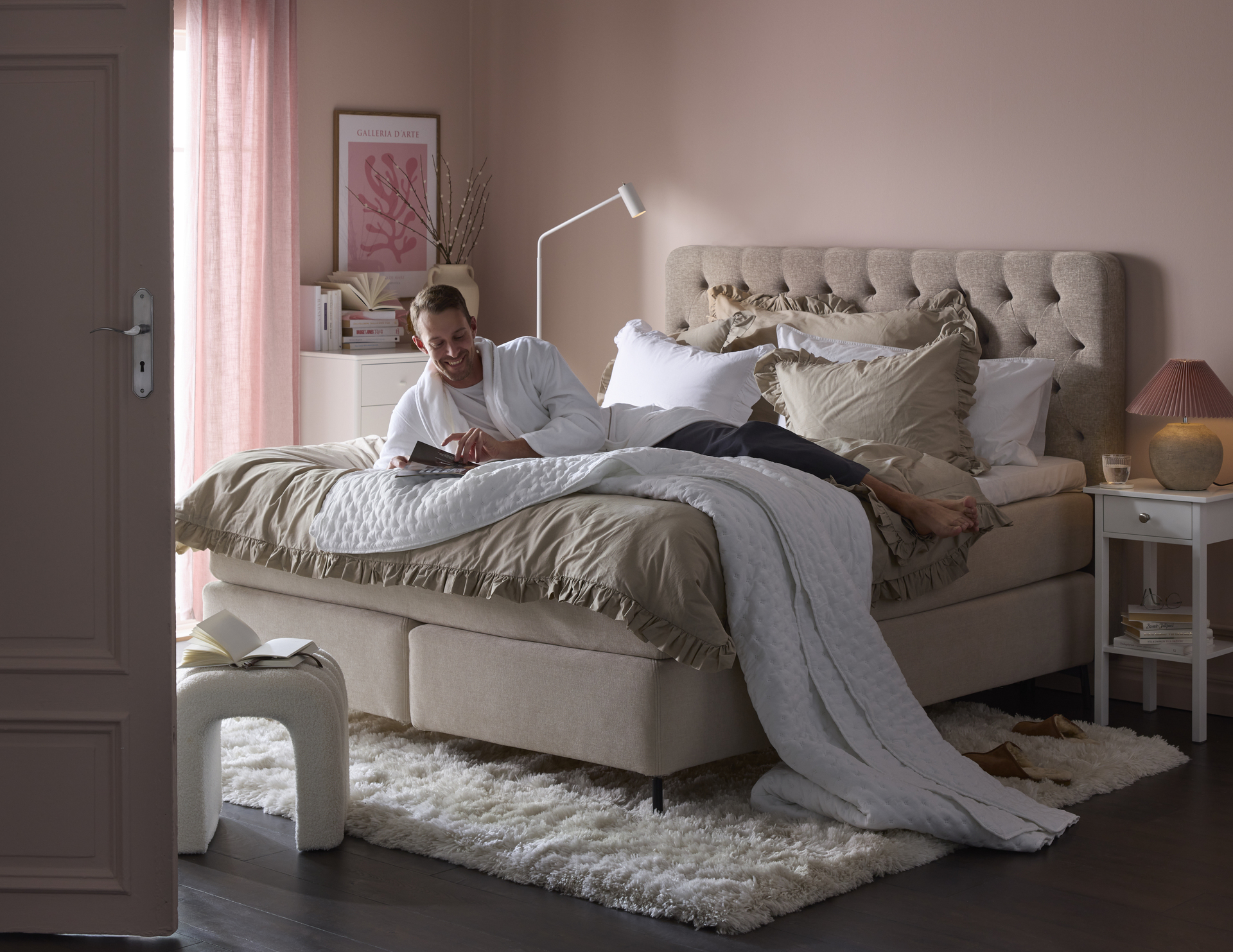 Ung man halvligger i en kontinentalsäng och bläddrar i magasin i ett rosaskimrande sovrum. 