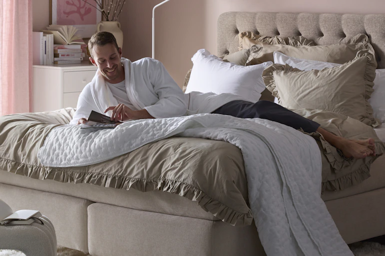 Ung man halvligger i en kontinentalsäng och bläddrar i magasin i ett rosaskimrande sovrum.