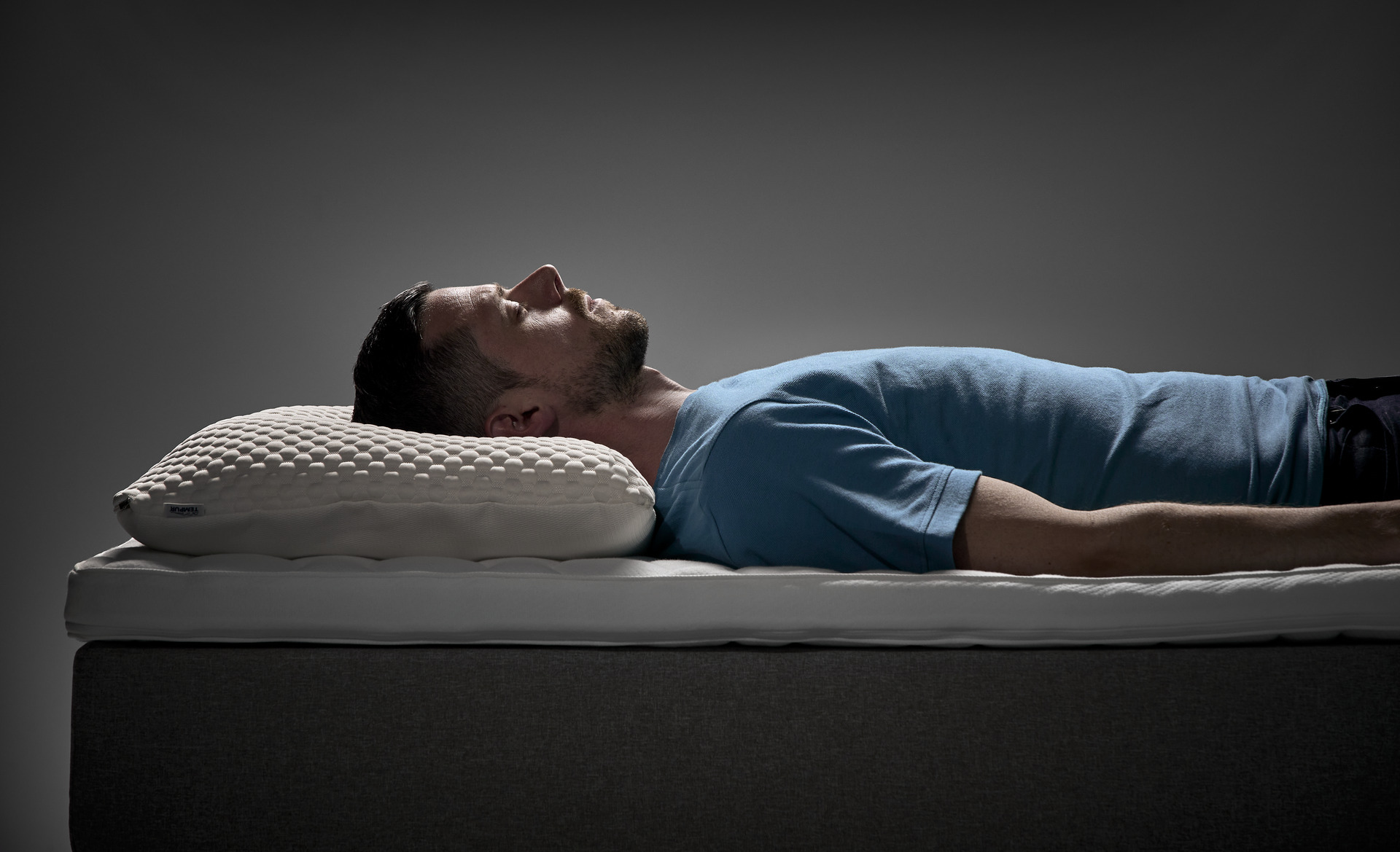 Man ligger på rygg i en säng med ergonomisk kudde under huvudet i en studiomiljö.