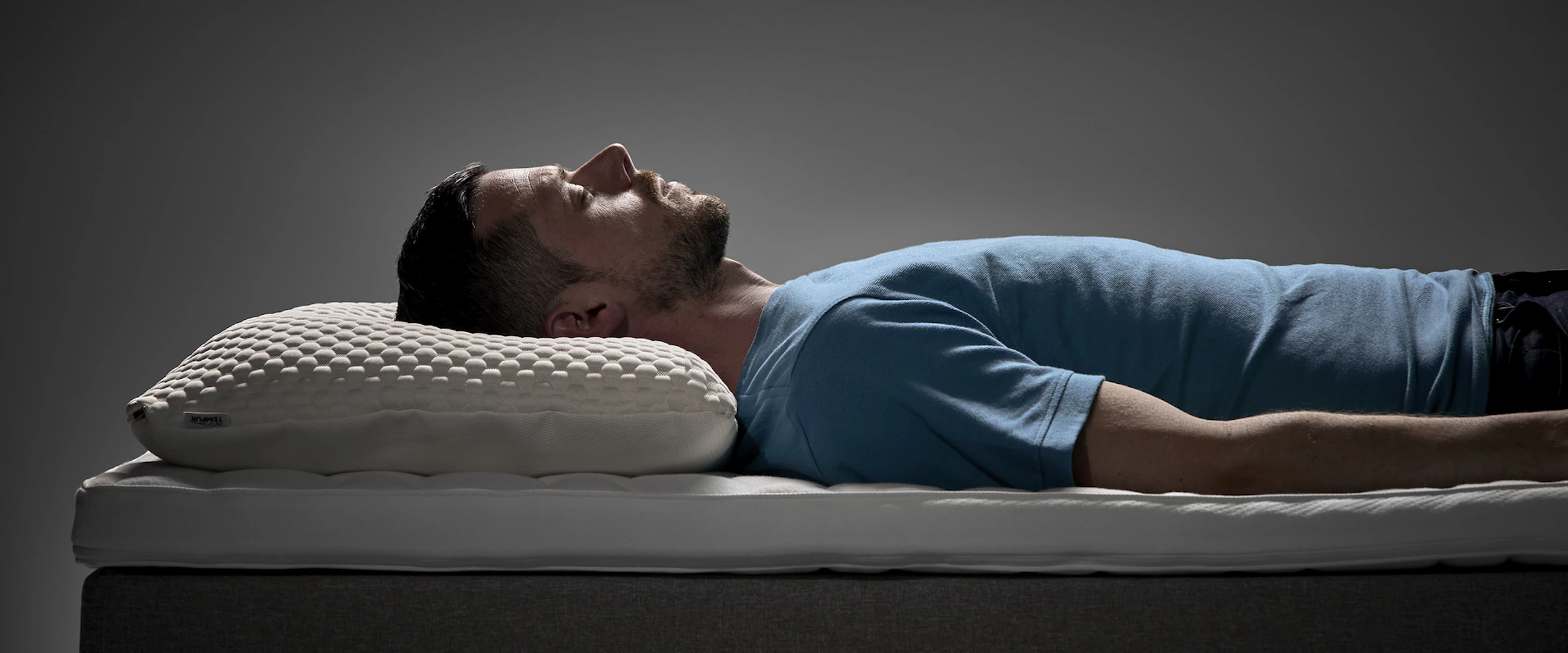 Man ligger på rygg i en säng med ergonomisk kudde under huvudet i en studiomiljö.