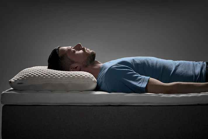 Man ligger på rygg i en säng med ergonomisk kudde under huvudet i en studiomiljö.