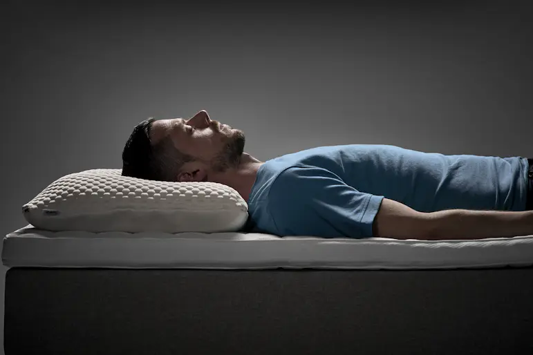 Man ligger på rygg i en säng med ergonomisk kudde under huvudet i en studiomiljö.