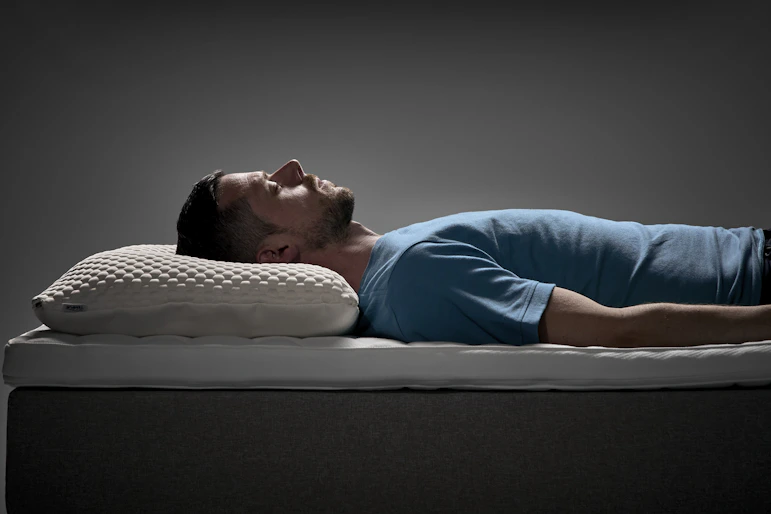 Man ligger på rygg i en säng med ergonomisk kudde under huvudet i en studiomiljö.