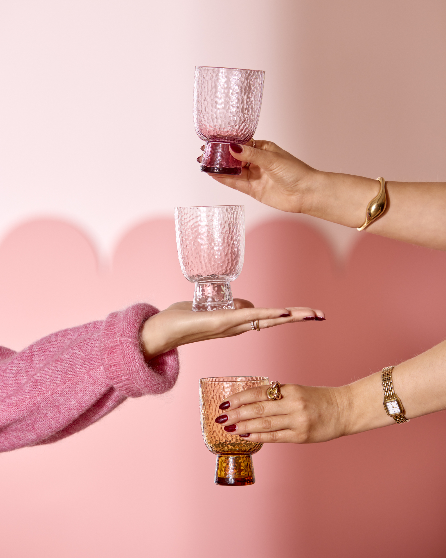 tre armar sticker fram och håller i tre olika glas framför en rosa vägg