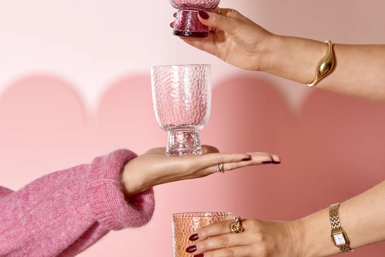 tre armar sticker fram och håller i tre olika glas framför en rosa vägg