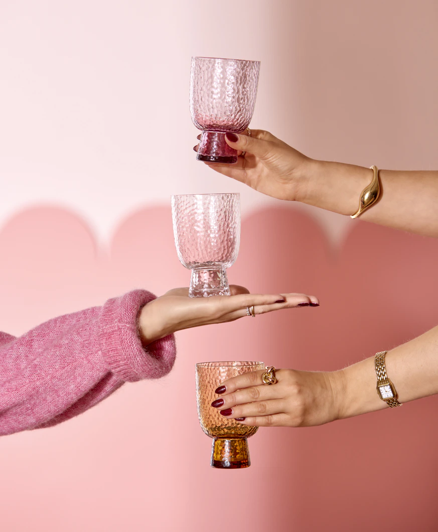 tre armar sticker fram och håller i tre olika glas framför en rosa vägg