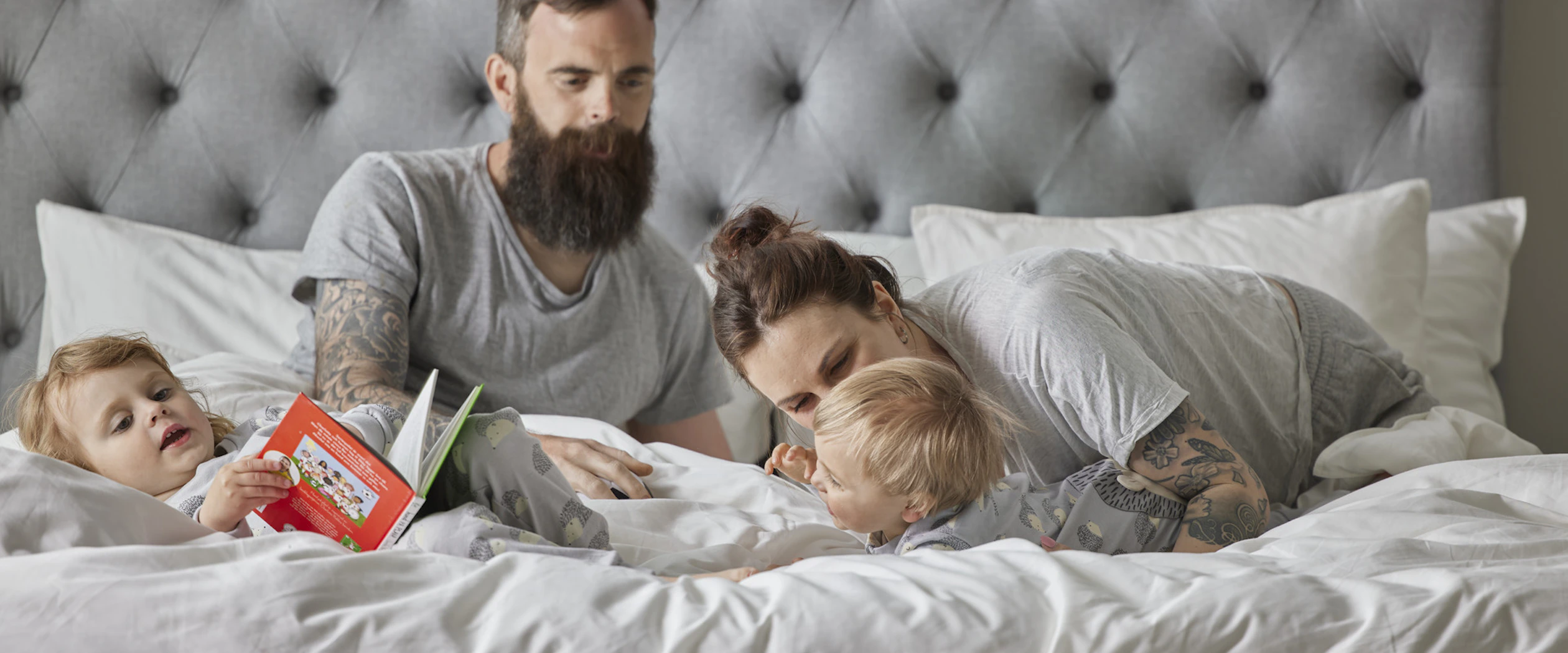 Familj med mamma, pappa och två små barn ligge roch myser i en ljusgrå säng.