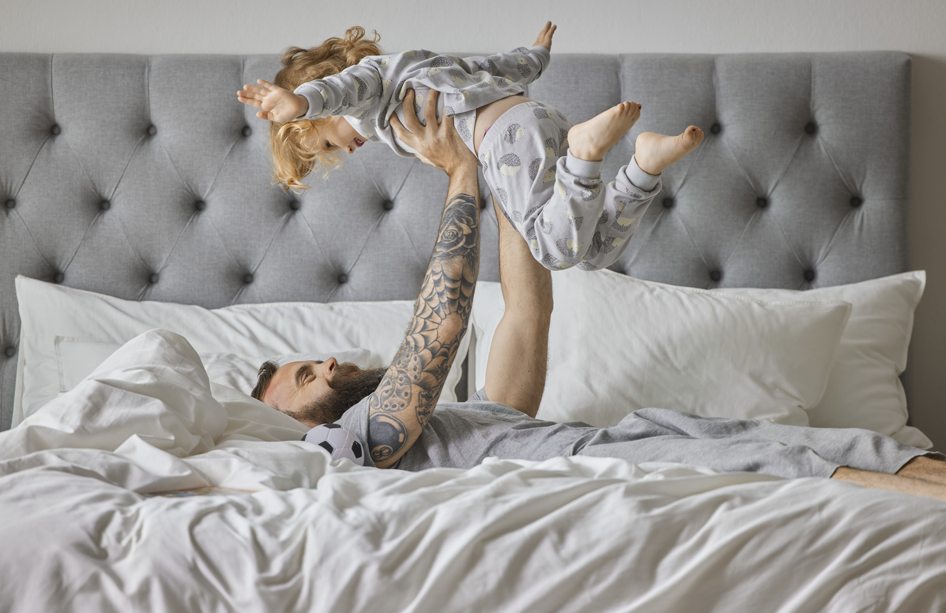 Pappa busar med sin dotter i en säng, ligger på rygg och håller upp henne och hon håller ut armarna som ett flygplan. 