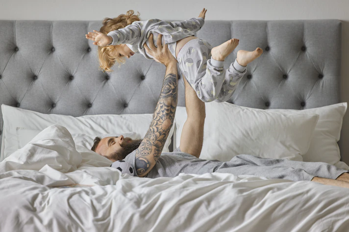 Pappa busar med sin dotter i en säng, ligger på rygg och håller upp henne och hon håller ut armarna som ett flygplan.