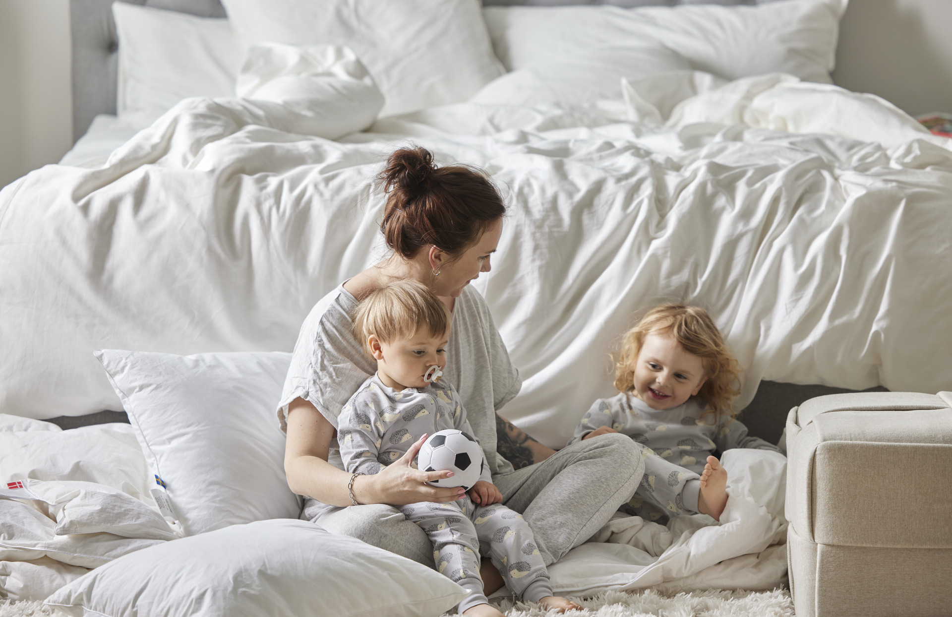 Mamma sitter på golvet nedanför säng och busar med sina två små barn.