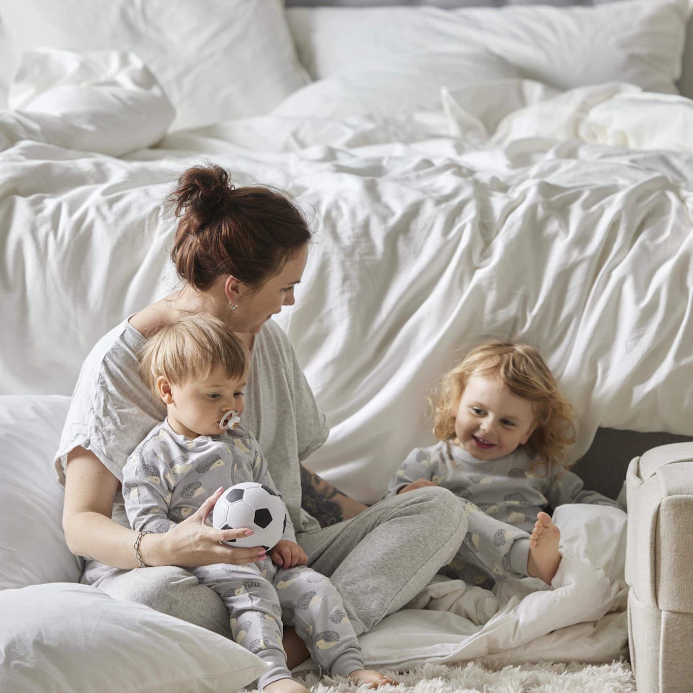 Mamma sitter på golvet nedanför säng och busar med sina två små barn.