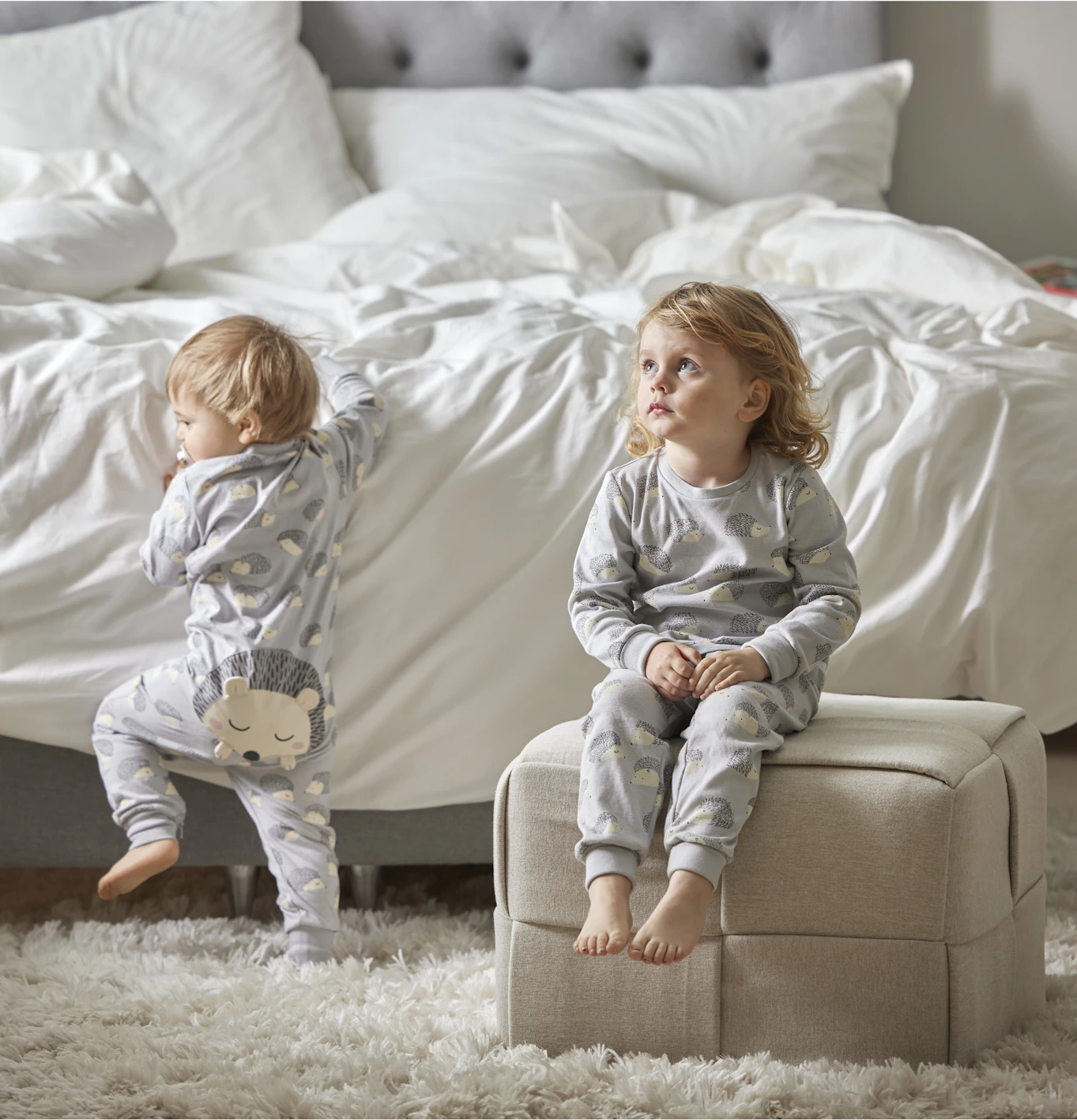 Två barn i sovrum, ett barn sitte rpå en puff framför sängen, det andra barnet som är yngre ser ut att försöka kliva upp på sängen. 