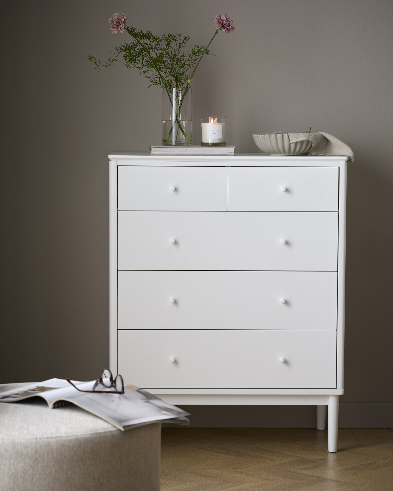 en vit byrå med en blomvas och skål står mot en grå vägg