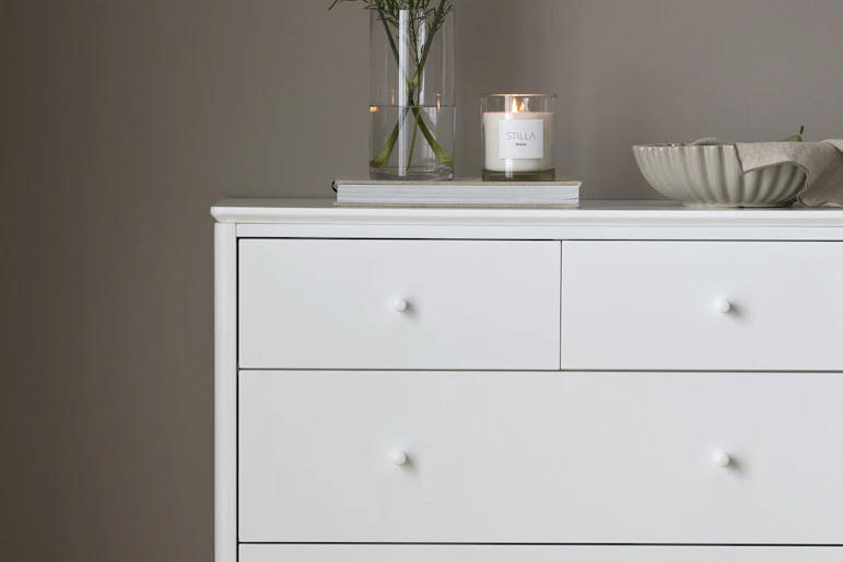 en vit byrå med en blomvas och skål står mot en grå vägg