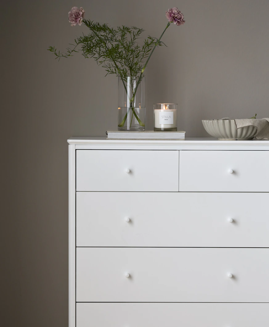 en vit byrå med en blomvas och skål står mot en grå vägg