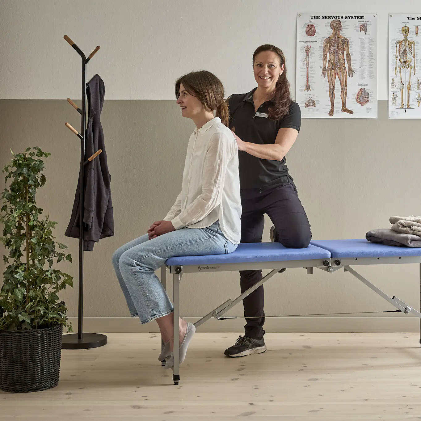 Monika Seth som är ergonom och fysioterapeut står bakom kund som sitter ned på kanten av en brits på en mottagning för fysioterapi, Monika känner på kundens rygg.