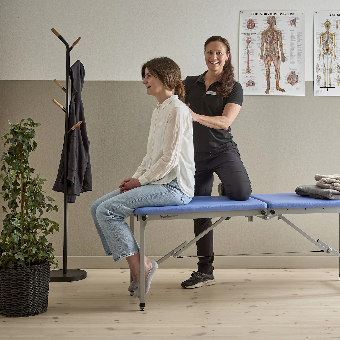 Monika Seth som är ergonom och fysioterapeut står bakom kund som sitter ned på kanten av en brits på en mottagning för fysioterapi, Monika känner på kundens rygg.