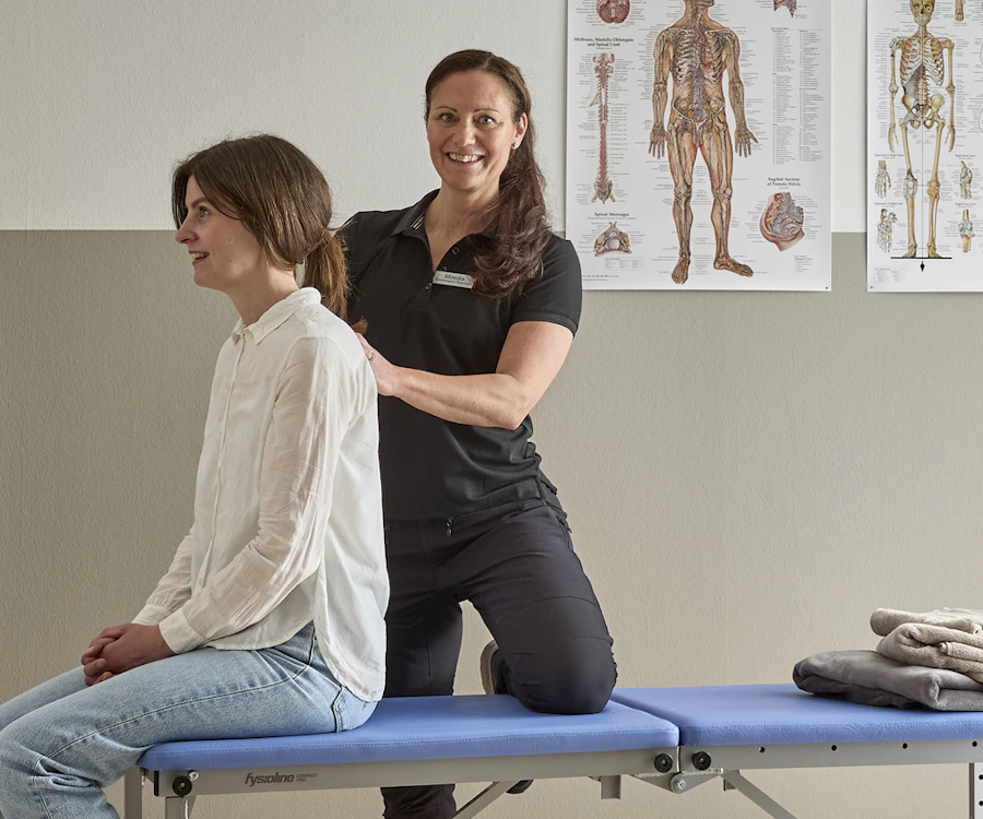 Monika Seth som är ergonom och fysioterapeut står bakom kund som sitter ned på kanten av en brits på en mottagning för fysioterapi, Monika känner på kundens rygg.