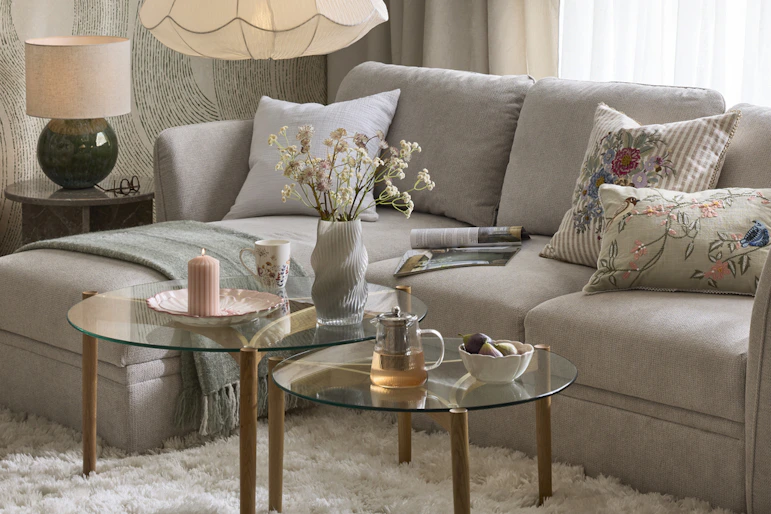 en grå bäddsoffa står i ett vardagsrum med en stor vit matta och soffbord i glas