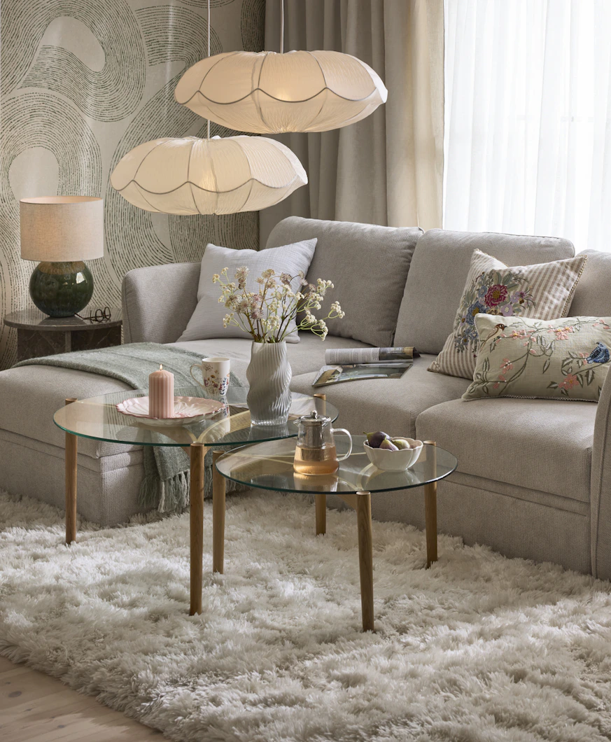 en grå bäddsoffa står i ett vardagsrum med en stor vit matta och soffbord i glas