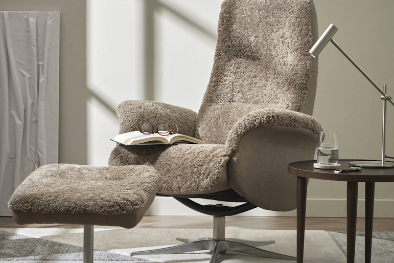 en grå fåtölj i fårskinn med fotpall står på en matta med ett sidobord bredvid