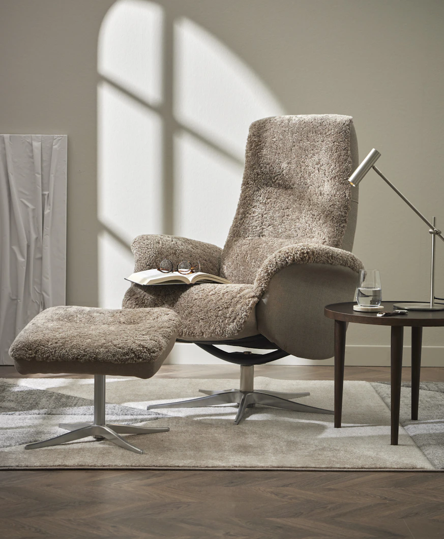 en grå fåtölj i fårskinn med fotpall står på en matta med ett sidobord bredvid