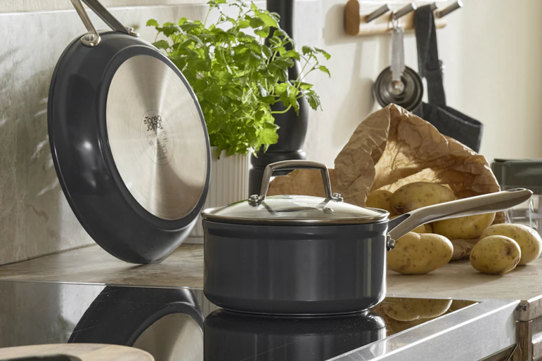 På en spis står en kastrull och på köksbänken bredvid står en mängd olika köksredskap