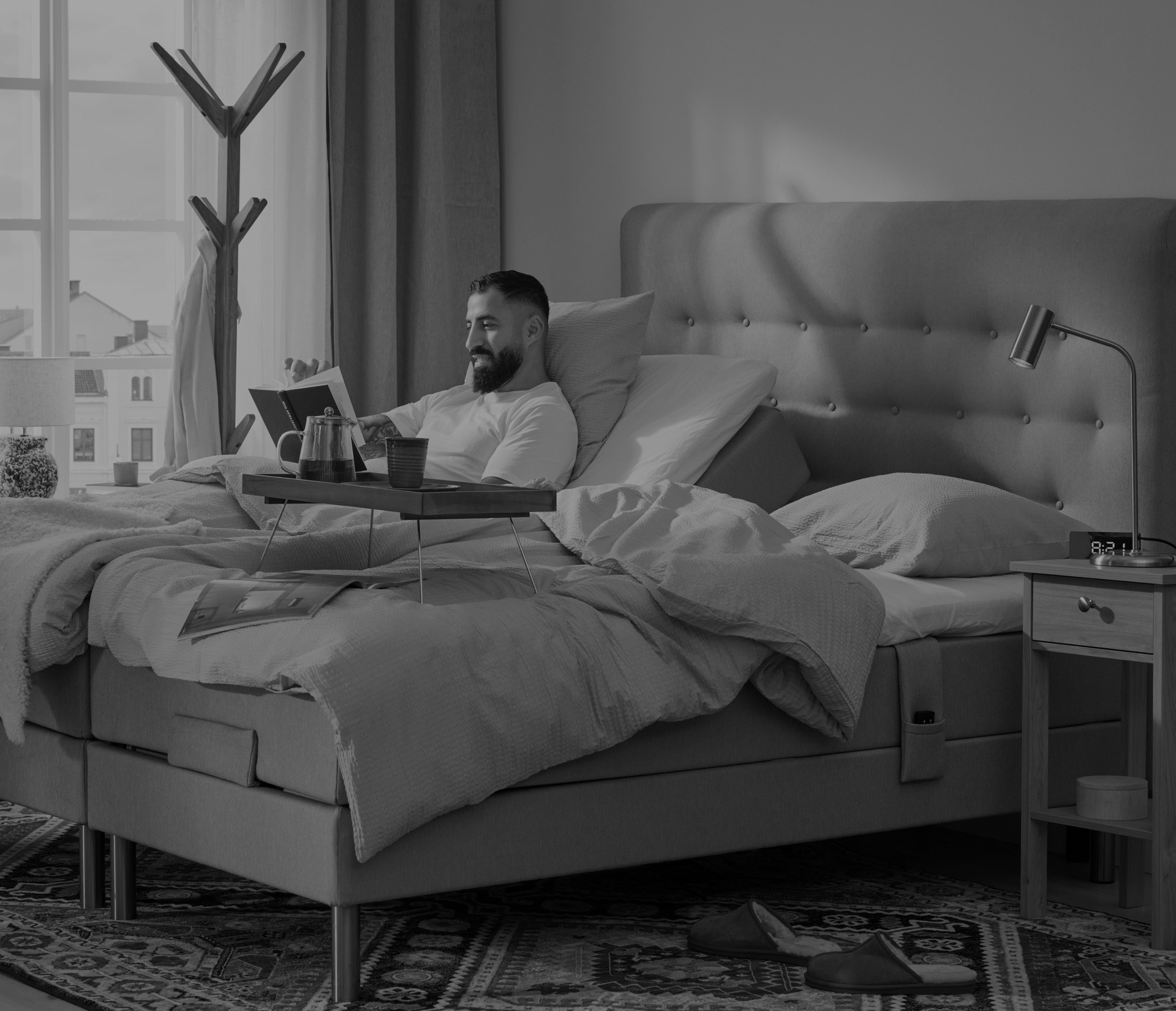 Man med skägg halvsitter upp och läser en bok i en ställbar kontintentalsäng i ett ljust sovrum där solen tittar in i fönstret. Bild i gråskala. 