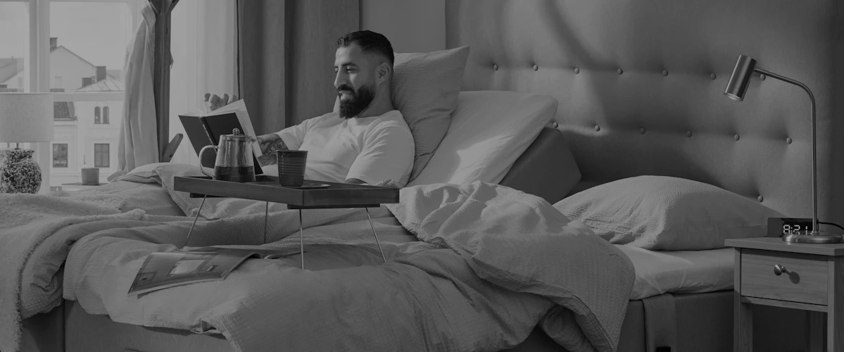 Man med skägg halvsitter upp och läser en bok i en ställbar kontintentalsäng i ett ljust sovrum där solen tittar in i fönstret. Bild i gråskala.