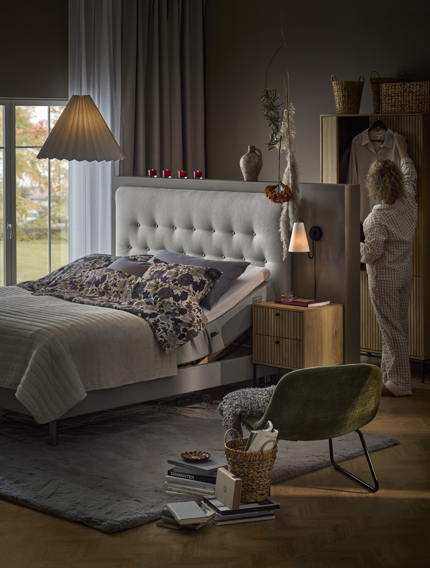 Sovrum med säng centrerade i mitten av rummet, kvinna i pyjamas står snett bakom sängen och hänger upp en galge med klänning i garderoben. 