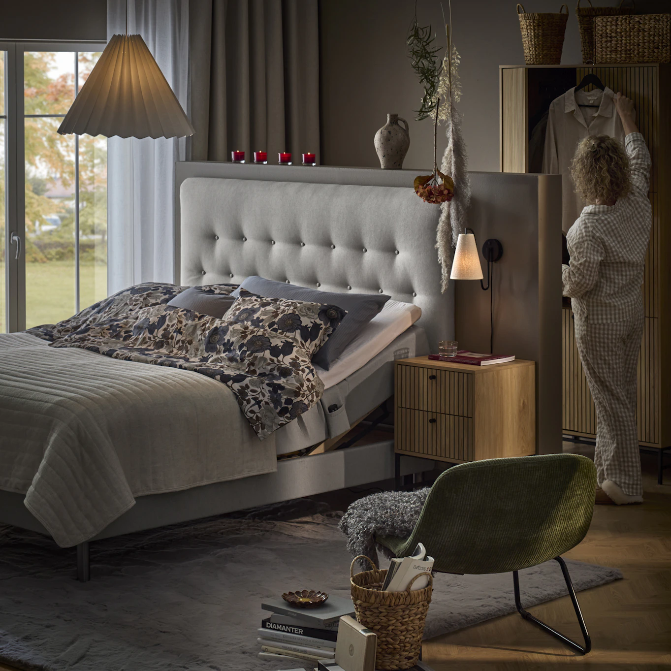 Sovrum med säng centrerade i mitten av rummet, kvinna i pyjamas står snett bakom sängen och hänger upp en galge med klänning i garderoben.