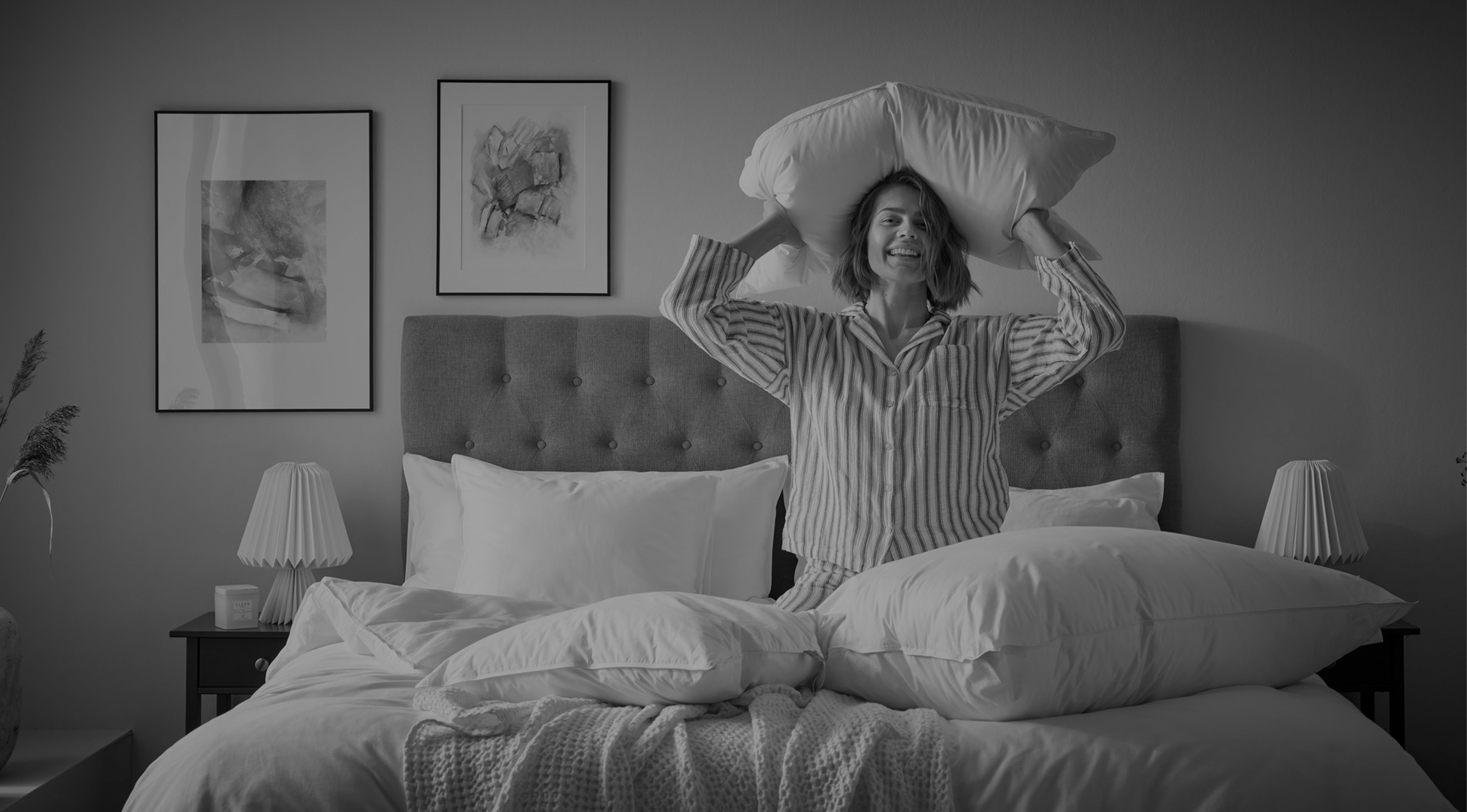Kvinna i randig pyjamas står på knä i en säng och ler mot kameran och håller en kudde bakom huvudet. Bild i gråskala.