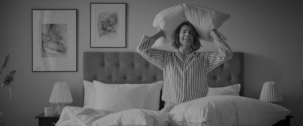 Kvinna i randig pyjamas står på knä i en säng och ler mot kameran och håller en kudde bakom huvudet. Bild i gråskala.